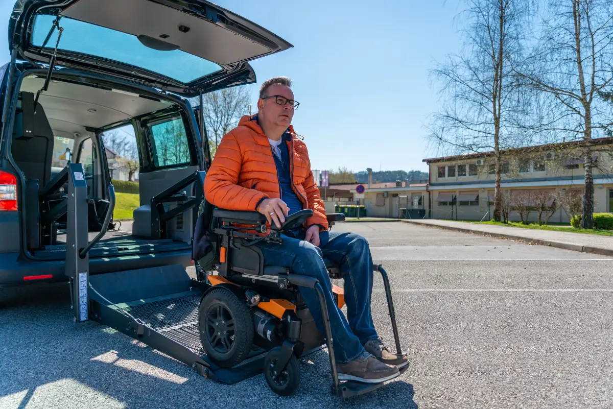 A man in an orange jacket sitting on the back of a wheelchair.