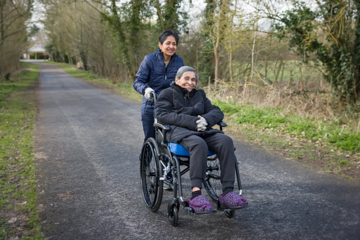 A woman pushing an elderly person in a wheelchair down the road.